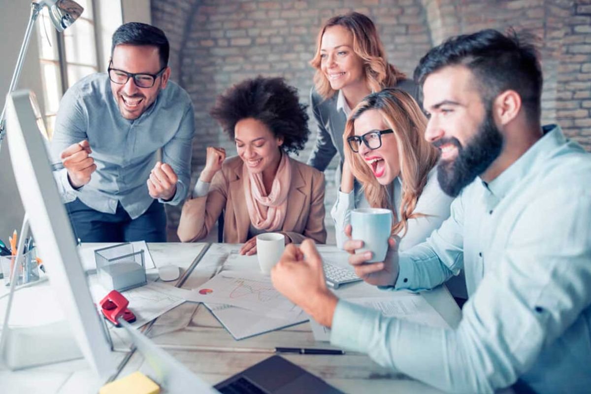 Um grupo de cinco colegas de trabalho, três homens e duas mulheres, estão reunidos em torno de uma mesa em um escritório moderno, todos sorrindo e comemorando enquanto olham para a tela de um computador. A cena transmite entusiasmo e sucesso, com papéis, canecas de café e materiais de escritório espalhados sobre a mesa. Todos parecem estar felizes e engajados, sugerindo um momento de vitória ou uma conquista importante no trabalho